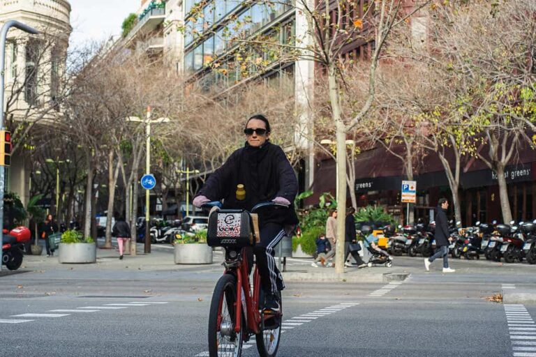 Un hombre sonriente con gafas pedaleando una bicicleta en una concurrida calle urbana, rodeado de árboles y edificios modernos, que representa un estilo de vida activo y ecológico.