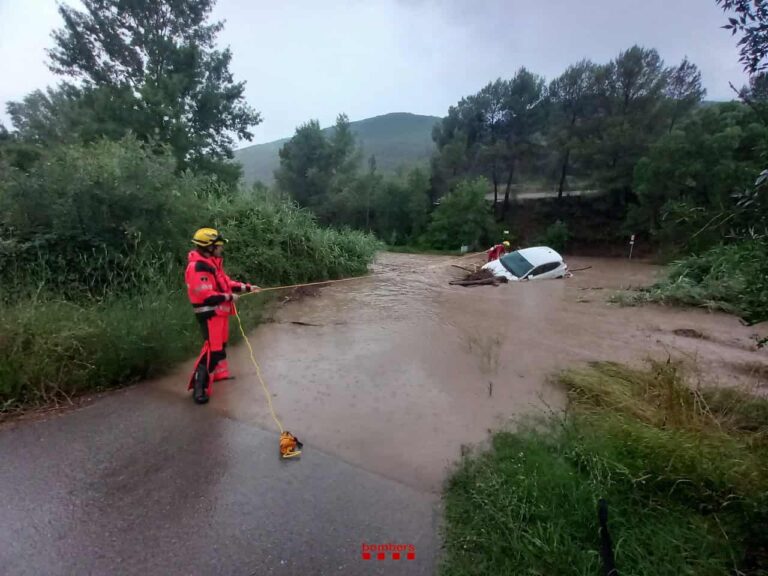Rescatista monitorizando una inundación en una carretera en Cataluña, con un coche parcialmente sumergido en el agua, rodeado de una densa vegetación y montañas al fondo.