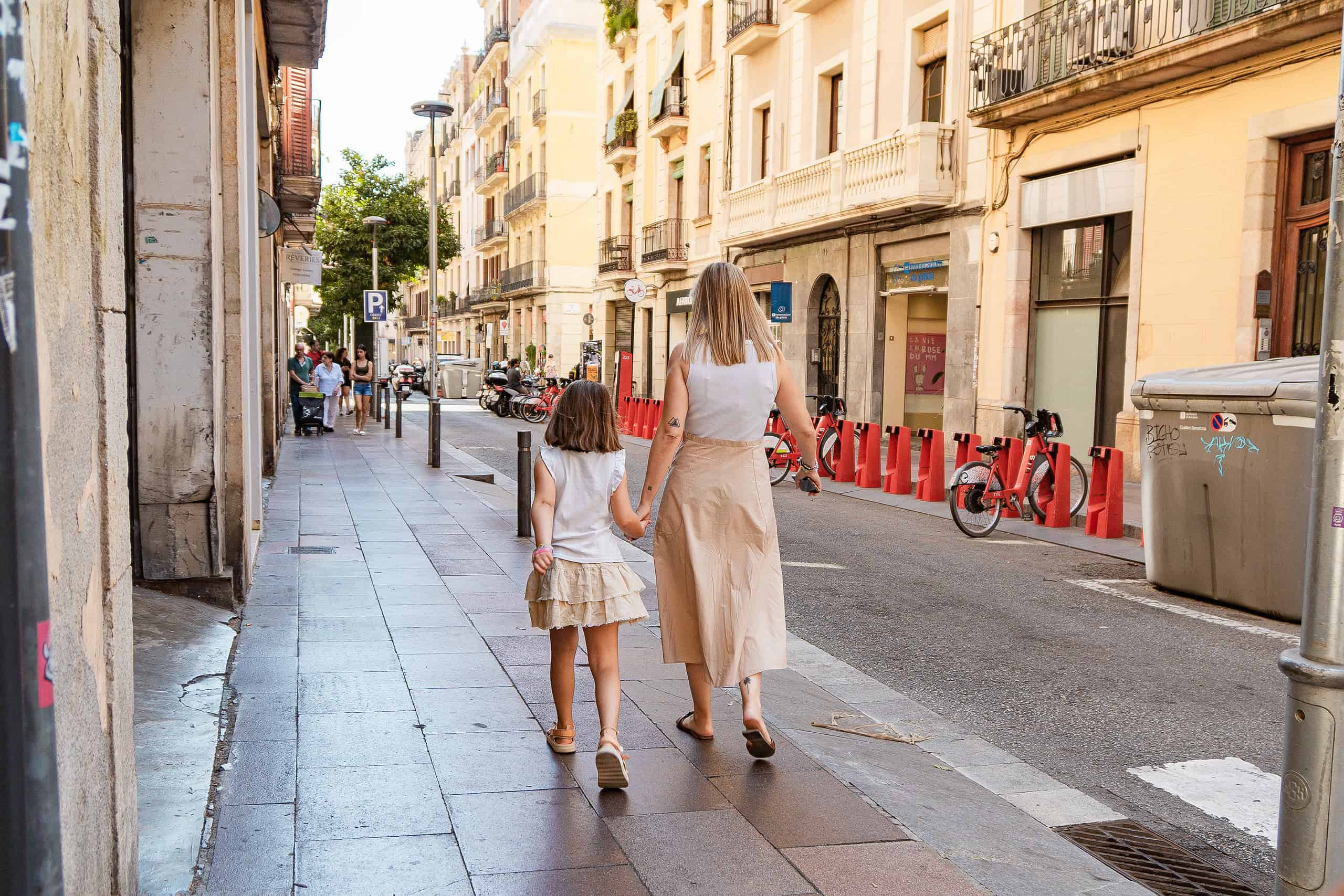 Madre e hija pasean por una concurrida calle de Barcelona, ​​con edificios históricos, bicicletas y gente pasando. Momento de complicidad y descubrimiento en un entorno urbano con encanto.