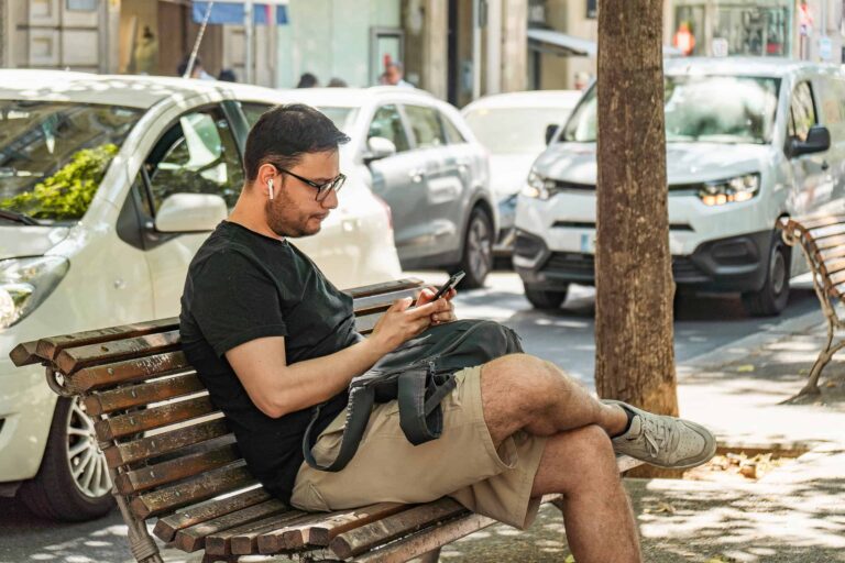 Hombre sentado en un banco de la ciudad, usando su teléfono inteligente, con autos al fondo.