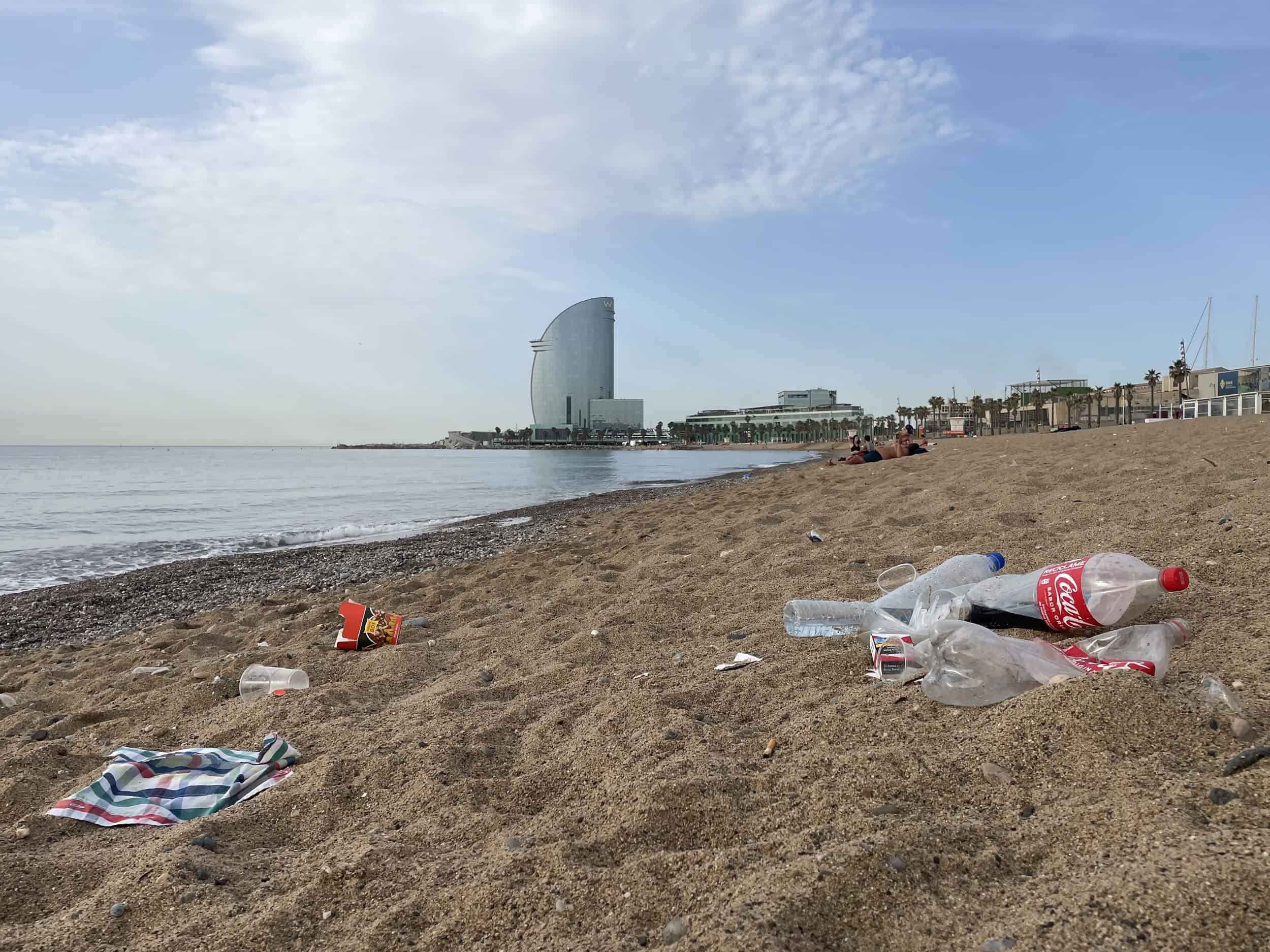 Barriendo residuos plásticos en la playa de Barcelona con la silueta del edificio W Barcelona al fondo, bajo un cielo parcialmente nublado.