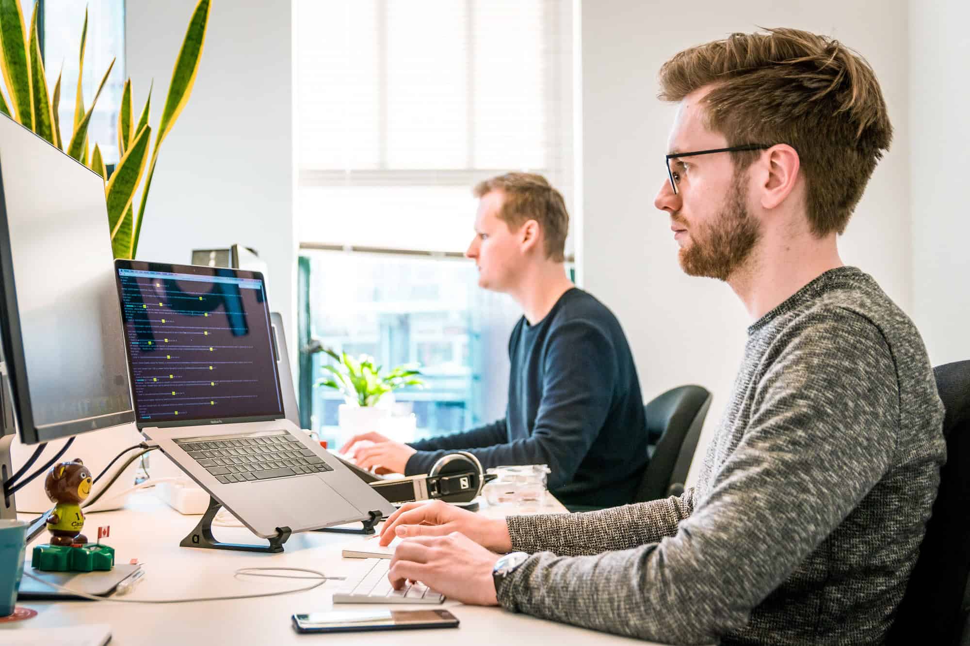 Por qué los salarios siguen tan bajos en España a pesar del crecimiento 2 Dos jóvenes trabajan en una oficina luminosa con computadoras, plantas y accesorios de oficina modernos, creando un ambiente de trabajo productivo e inspirador.