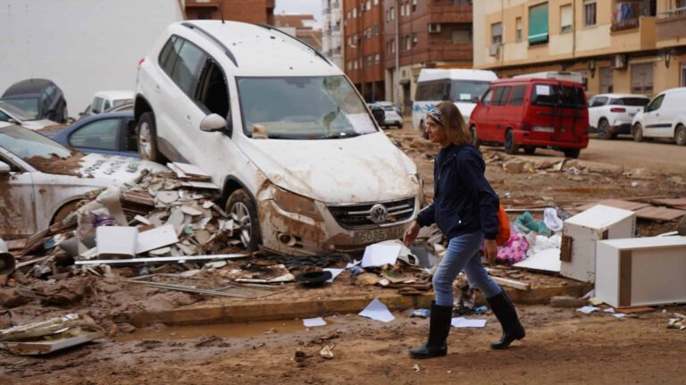 Mujer caminando entre un paisaje de escombros y vehículos dañados tras las inundaciones en Valencia, ilustrando la destrucción causada por el desastre natural.