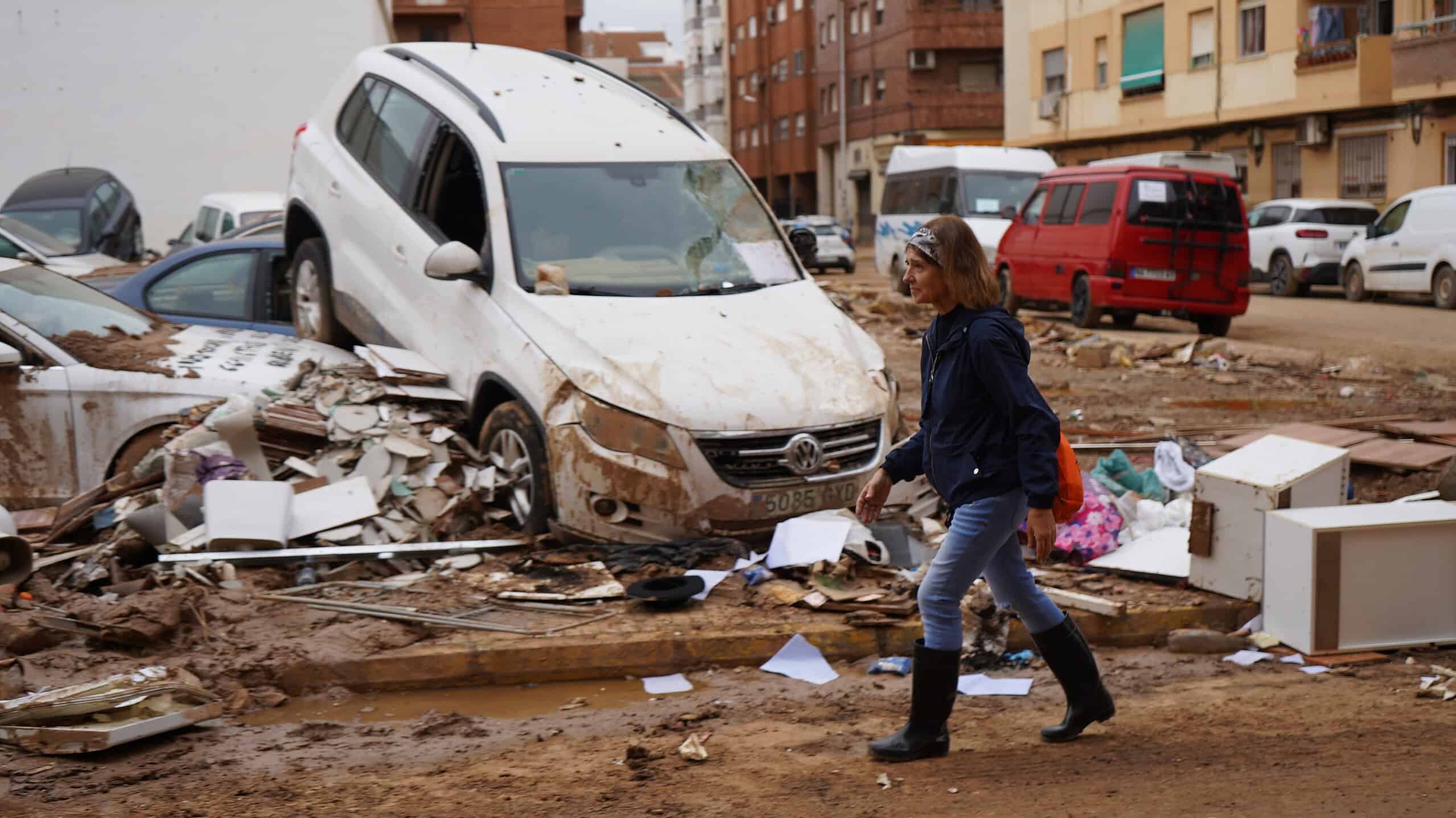 Mujer caminando entre un paisaje de escombros y vehículos dañados tras las inundaciones en Valencia, ilustrando la destrucción causada por el desastre natural.